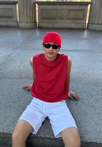 Κόκκινο χωρίς μανίκια τοπ και κόκκινο καπέλο με μαύρα γυαλιά ηλίου. Λευκές σορτς. Καθισμένος σε μια πέτρινη επιφάνεια με υφή γκρι φόντο.