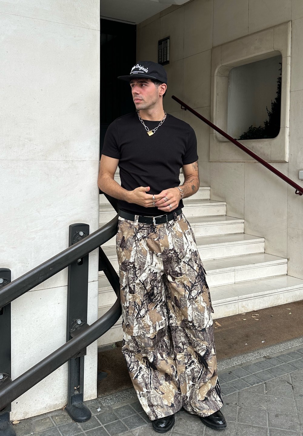 T-shirt ajustado preto, calças cargo camufladas com padrões castanho, bege e preto, cinto preto, corrente prateada, boné preto e botas.