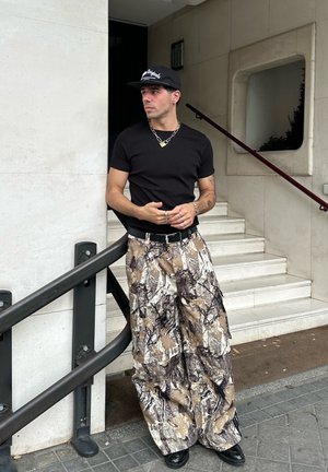 Camiseta ajustada negra, pantalones cargo camuflados con patrones en marrón, beige y negro, cinturón negro, collar de cadena plateada, gorra negra y botas.