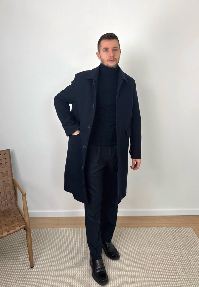 Man standing indoors wearing a black overcoat, turtleneck, trousers, and leather shoes, with one hand in his coat pocket.
