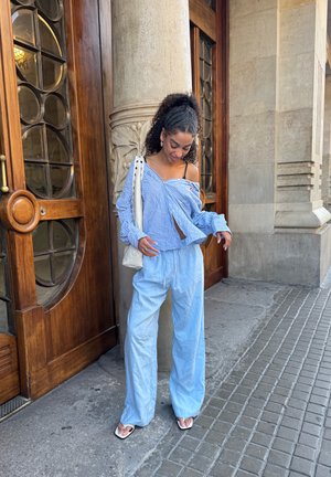 Camisa de rayas azul y blanca, pantalones anchos de un azul claro y sandalias blancas. Sosteniendo una bolsa de tela color crema, de pie cerca de puertas de madera.