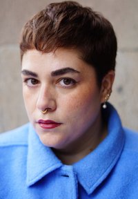 Lühikeste pruunide juuste, pruuni silmavärviga inimene, punase huulepulgaga, kuldse septum-rõnga ja mitme kuldse kõrvarõngaga, sinist mantlit kandes, vaatab kaamerasse.