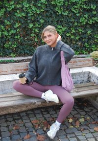Grijze geribbelde trui met hoge hals, roze legging, witte sneakers. Zittend op een bankje met een koffiekop in de hand en een roze schoudertas.