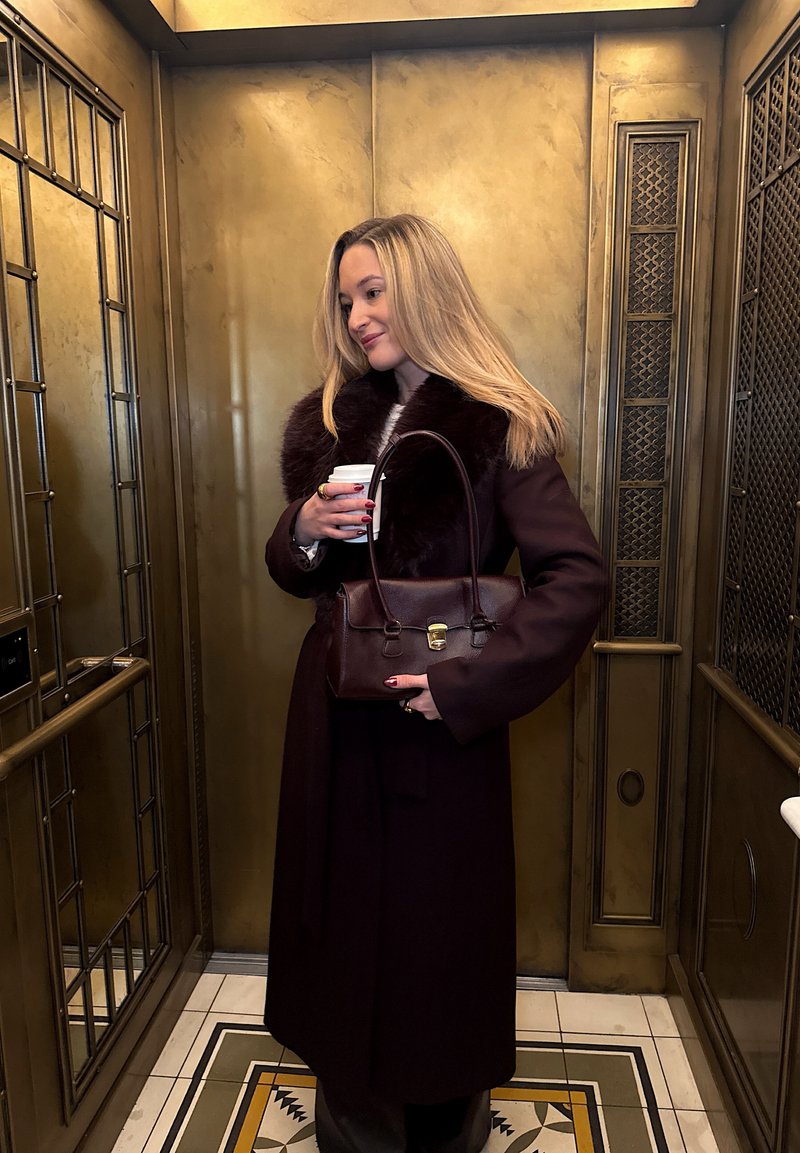 Femme blonde portant un long manteau foncé à col en fourrure tient une tasse de café et un sac à main en cuir assorti dans un ascenseur en laiton vintage avec un sol à motifs.