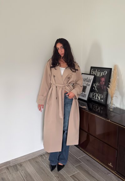 Abrigo trench beige con cinturón, diseño de doble botonadura y grandes solapas. Lleva una blusa blanca debajo y lo combina con jeans azules y tacones negros.