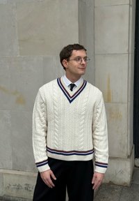 Krem pulover z žepi z vzorcem pletenja v obliki kablov, z globokim V-izrezom, poudarjen z temno modrimi in bordo črtami na ovratniku in spodnjem robu, nošen čez belo srajco.