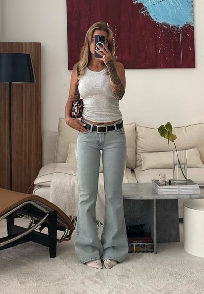 Camiseta sin mangas gris, jeans de campana azul claro, cinturón negro con hebilla plateada y bolso de estampado de leopardo. La habitación cuenta con un sofá beige y una mesa gris.