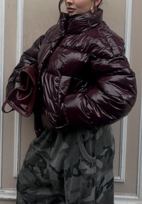 Burgundy padded jacket with high collar, shiny finish, and button fastening. Grey camo trousers with drawstring. Holding a matching handbag.