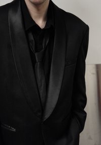 Veste de smoking noire avec revers en satin, assortie à une cravate noire et une chemise noires. Tissu lisse, coupe ajustée, avec une poche latérale.