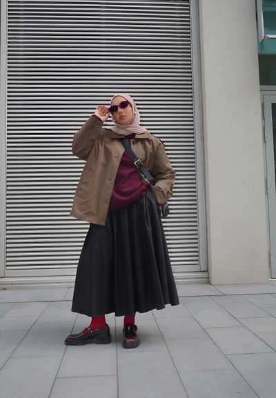 Chaqueta marrón de gran tamaño, suéter burdeos, falda negra de corte completo, calcetines rojos y zapatos negros con acentos rojos. Las gafas de sol añaden un toque moderno.