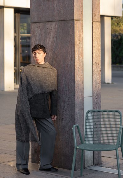 Persona con cabello corto y rizado, vistiendo un blazer oscuro y una larga bufanda, apoyada en una columna de piedra cerca de una silla verde vacía al aire libre.