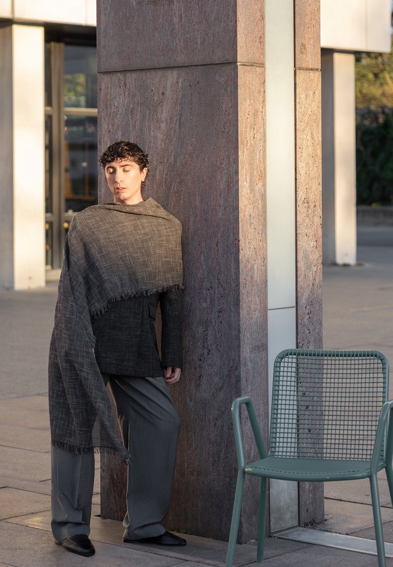 Personne aux cheveux courts et bouclés, vêtue d'un blazer foncé et d'une longue écharpe, appuyée contre un pilier en pierre près d'une chaise verte vide à l'extérieur.