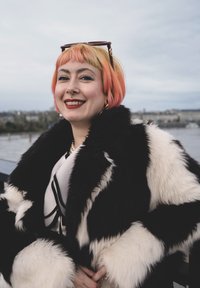 Svart-hvitt fuskepelsfrakk med fluffy tekstur og kontrasterende paneler. Personen har kort, flerfarget hår og store øredobber.