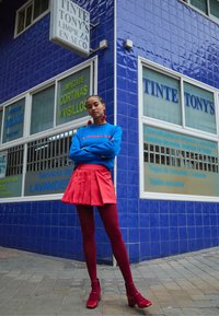 Sweatshirt azul com logótipo vermelho, saia plissada vermelha e meias combinando. Sapatos de plataforma vermelhos. O fundo apresenta uma parede de azulejos azuis e sinalização da loja.