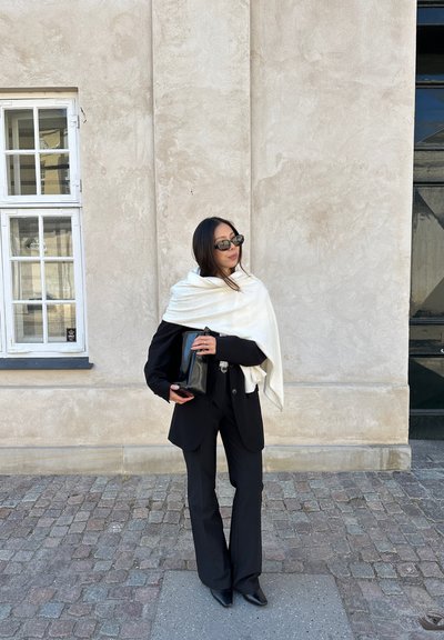 Blazer negro entallado y pantalones acampanados con un chal blanco. Sosteniendo un clutch negro, usando gafas de sol negras frente a una pared texturizada.