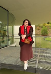 Brun kavaj över en beige topp, röd scarf, brun kjol och sneakers. En ljusbrun väska vilar mot kroppen, med en suddig grön bakgrund.