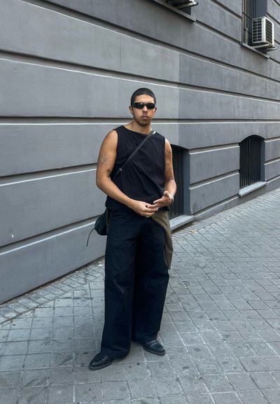 Top negro sin mangas y pantalones anchos negros, de pie en una calle pavimentada. Los accesorios incluyen gafas de sol y un bolso de hombro con un patrón de cuadros.