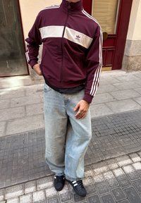 Jachetă cu fermoar în nuanță de burgundă, cu o dunga orizontală albă, purtată cu blugi evazați de culoare albastră deschisă. Tenisi negri cu dungi albe. Outfit urban casual.