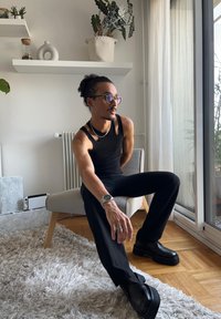 Haut sans manches noir avec des détails découpés, pantalon noir à large jambe et chaussures noires imposantes. Lunettes claires et colliers en argent en couches. Chaise de couleur claire.