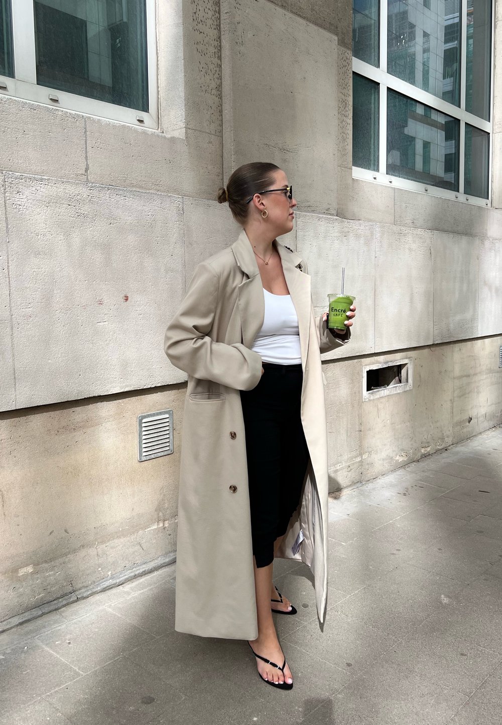 Femeie îmbrăcată într-o haină lungă bej, pantaloni negri scurți, bluză albă și sandale, ținând un smoothie verde lângă o clădire din piatră cu feronerie.