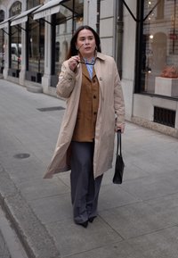 Beige trenchcoat, bruine vest, lichtblauwe shirt, grijze broek, houdt een zonnebril vast, zwarte tas, stedelijke omgeving met winkels op de achtergrond.