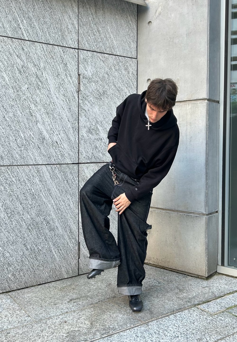 Svart huva sweatshirt, lösa mörka denimjeans med uppvikta fållar, metallisk detaljer, står på en strukturerad stonyta.