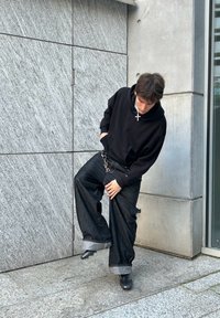 Svart huva sweatshirt, lösa mörka denimjeans med uppvikta fållar, metallisk detaljer, står på en strukturerad stonyta.