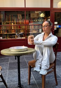 Hvit treningsdrakt med glidelås, båret av en sittende person som holder et glass med sjokoladedrikk, med et rundt metallfat på et trebord. Bakgrunnen har hyller med fargerike bøker.