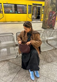 Bruine suède jas, zwarte maxi jurk, gevlochten haar en blauwe schoenen. Houdt een geweven bordeauxrood handtas vast in een metrostation.