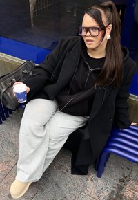 Sort jakke over en sort hættetrøje, grå joggingbukser og beige sko. Sidder på en blå bænk med en kaffekop i hånden og en grå håndtaske i nærheden.