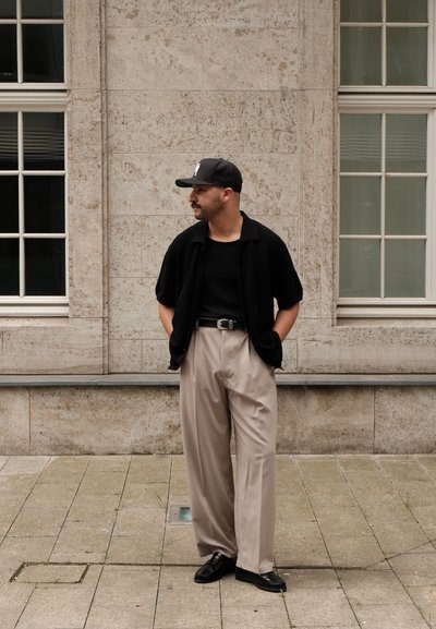 Cárdigan corto de punto negro con camiseta negra, pantalones beige de pinzas, zapatos de cuero negros y una gorra oscura de béisbol. Fondo de pared de piedra.