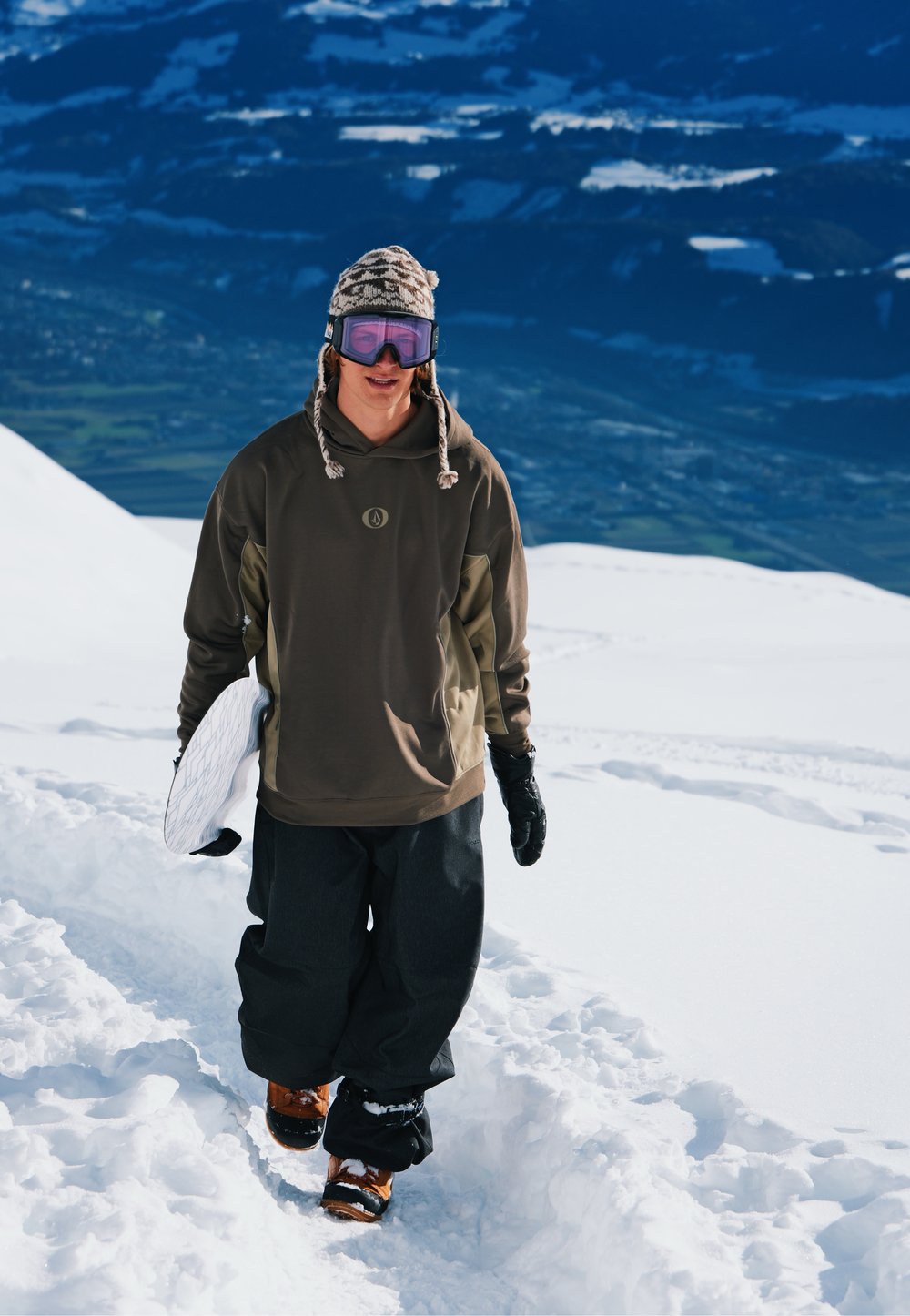 Snowboarder portant une veste marron à capuche et un pantalon noir, marchant sur la neige avec des bottes marron et un snowboard blanc sous le bras ; arrière-plan enneigé.
