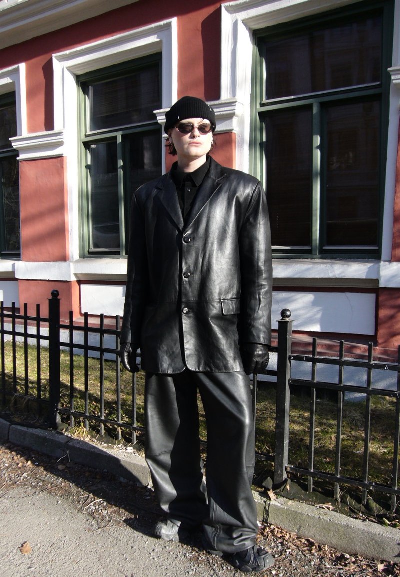 Άτομο που φοράει υπερμεγέθη μαύρο δερμάτινο μπουφάν και παντελόνια, μαύρο σκούφο, γάντια και γυαλιά ηλίου, στέκεται δίπλα σε φράχτη και κόκκινο κτίριο με πράσινα παράθυρα.