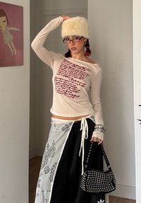 Chemise beige à manches longues avec texte rouge, associée à une jupe noire à cordon avec des accents en dentelle blanche. Sac à main noir clouté et chapeau en fausse fourrure.