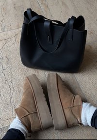 Bottines en daim marron avec semelles épaisses portées avec des chaussettes blanches, posées sur un sol carrelé près d'un sac à main en cuir noir.