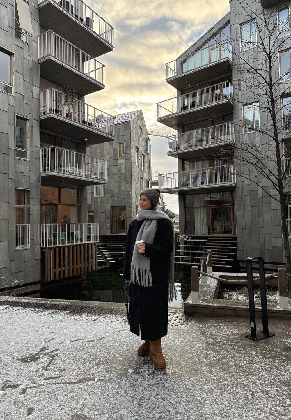 Edifícios de apartamentos modernos e cinzentos com varandas. Uma pessoa com um longo casaco preto e um cachecol cinzento segura uma chávena de café, em pé sobre uma superfície coberta de neve.