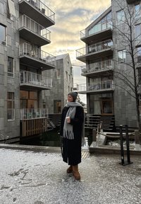 Edifícios de apartamentos modernos e cinzentos com varandas. Uma pessoa com um longo casaco preto e um cachecol cinzento segura uma chávena de café, em pé sobre uma superfície coberta de neve.