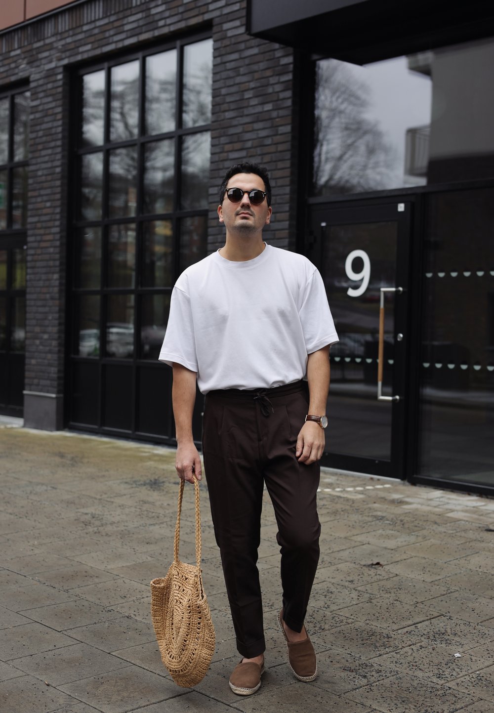 Vit oversized t-shirt, bruna tapered byxor, vävd tan väska, solglasögon, klocka. Stilig streetstyle-look mot en vägg av tegel.