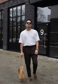Vit oversized t-shirt, bruna tapered byxor, vävd tan väska, solglasögon, klocka. Stilig streetstyle-look mot en vägg av tegel.