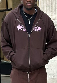 Brun zip-up hoodie med pink stjernegrafik på brystet, store lommer og en løs pasform. Matchet med brune bukser.