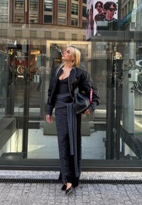 Sort bomberjakke over en sort bodycon-top, parret med en tekstureret, lang sort nederdel. Holder en sort taske, står ved siden af en glasbutik.