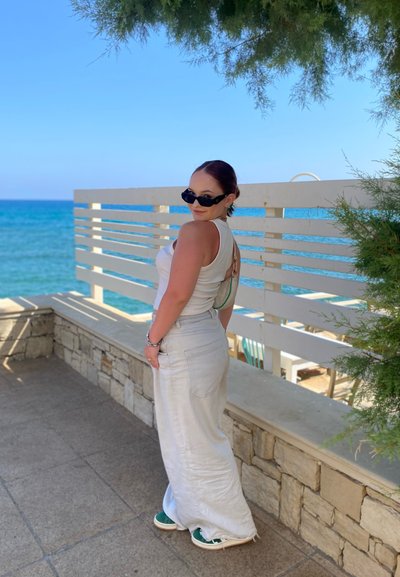 Top blanco sin mangas con la espalda descubierta, falda larga de color claro y zapatillas verdes. El fondo presenta una pared de piedra y vista al océano.