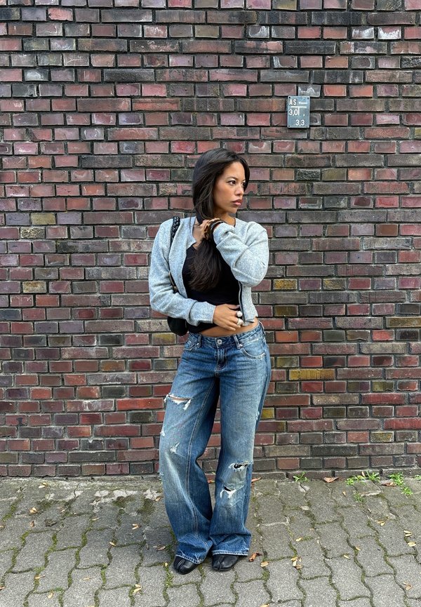 Femme aux longs cheveux foncés portant une veste grise, un haut noir et un jean bleu déchiré, se tenant dans une rue pavée contre un mur en briques rouges.