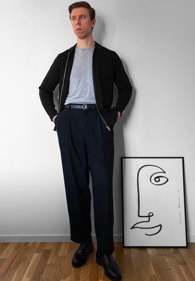 Chaqueta negra con cremallera sobre una camiseta gris, combinada con pantalones plegados azul marino y zapatos negros. Hay cerca una obra de arte en dibujo lineal.