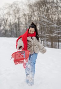 Jeune femme en manteau de fourrure et écharpe rouge, donnant un coup de pied dans la neige tout en tenant un grand sac à main rouge dans un décor hivernal avec des arbres dénudés.