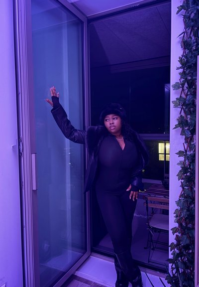 Mono negro con mangas largas, detalles de piel sintética y botas hasta la rodilla, frente a una puerta de cristal de tonos morados y vegetación.