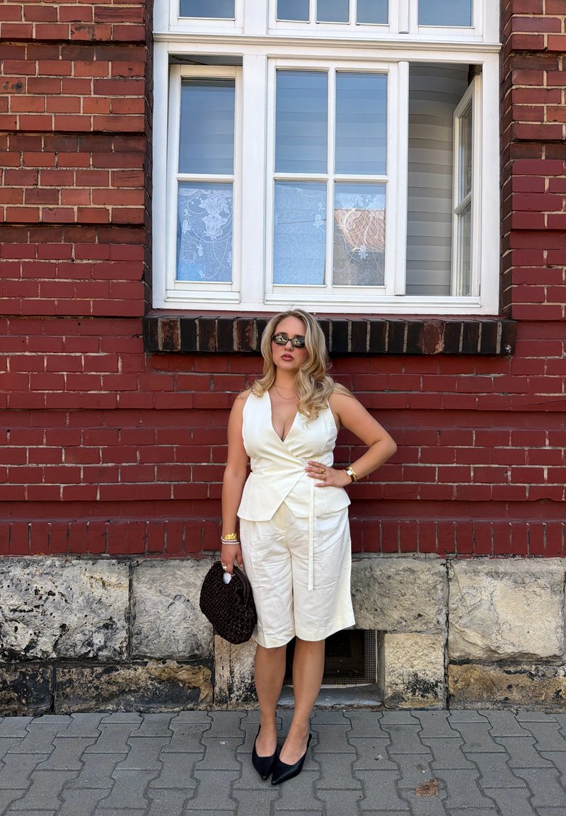 Femme en haut blanc sans manches et short avec des lunettes de soleil foncées et des chaussures noires pointues se tient contre un mur en briques rouges en tenant un sac à main tissé foncé.