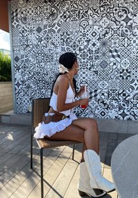 Valkoinen röyhelöinen toppi ja hame, ruskea vyö, jossa on pyöreitä yksityiskohtia, valkoiset cowboy-kengät, juoma kädessä, istuu musta-valkoista kuvioitua seinää vasten.