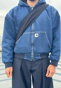 Tumši zila džinsu jaka ar rāvējslēdzēju priekša, kapuci un elastīgām piedurknēm. Iekļauj logo uzlīmi un valkāta ar melniem biksēm un plecu somu.