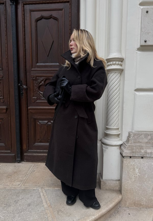 Lange, donkerbruine wollen jas met een hoge kraag, knoopsluiting en brede mouwen, gecombineerd met zwarte handschoenen en broek.